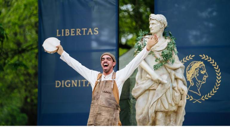 Acteur in uitbundige pose voor standbeeld van Julius Caesar. Op de achtergrond blauwe banieren met tekst in het Latijn