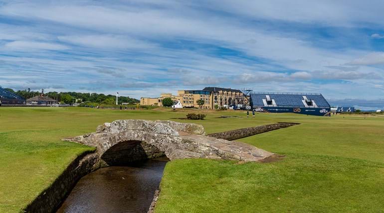 Terrain de golf traversé par un fossé et un vieux pont de pierre avec de grands bâtiments en arrière-plan 