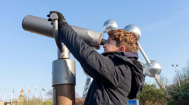 A teenage boy looks through a giant telescope. In the background the Brussels Atomium