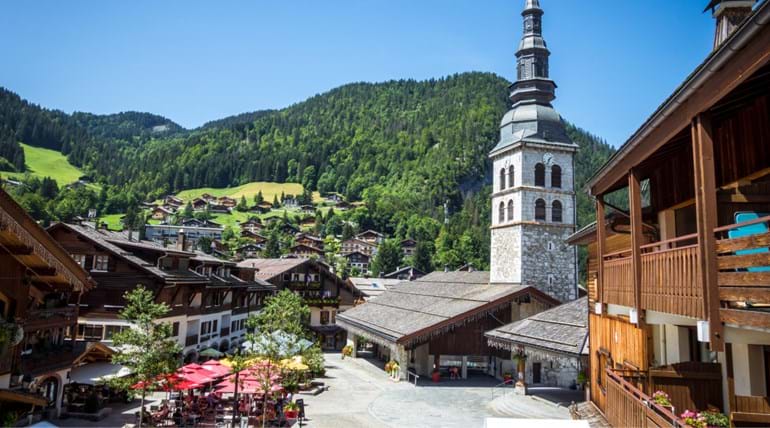 A large bell tower of a church dominates a town square with Alpine type buildings around it, on a summer’s day