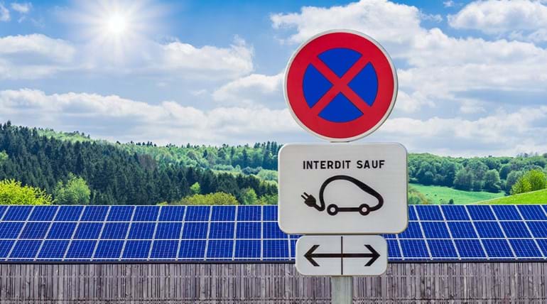 A French parking sign with an electric car symbol and a warning roundel sign, in front of a row of solar panels and a wooded area behind it