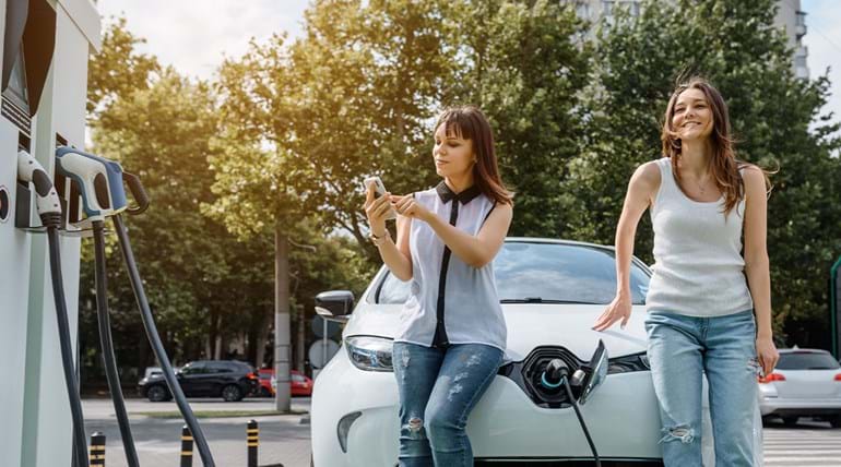 Two young women standing at the front of their electric car while it charges up, with one using her phone to pay