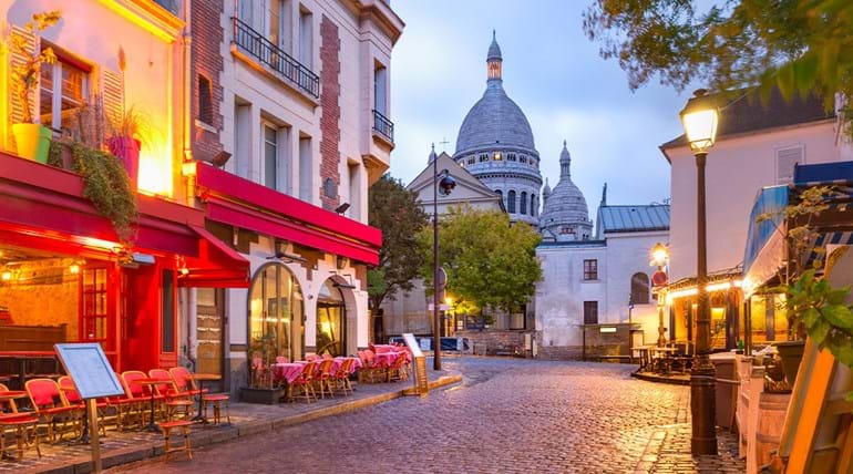 Pavement restaurants on a cobbled street at dusk, with the dome of a church in the background