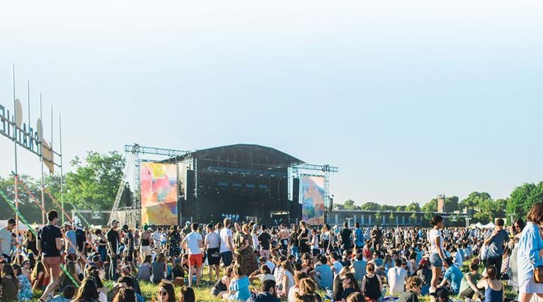 A main stage area at a festival, with music fans congregated in a field in front of a large stage 