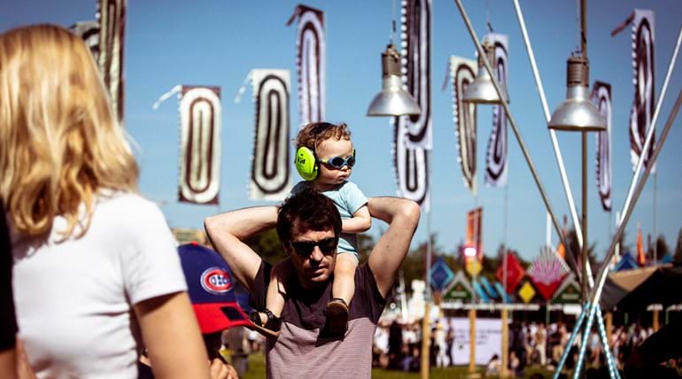 A toddler with large earplugs in is sitting on his father’s shoulders at a festival 